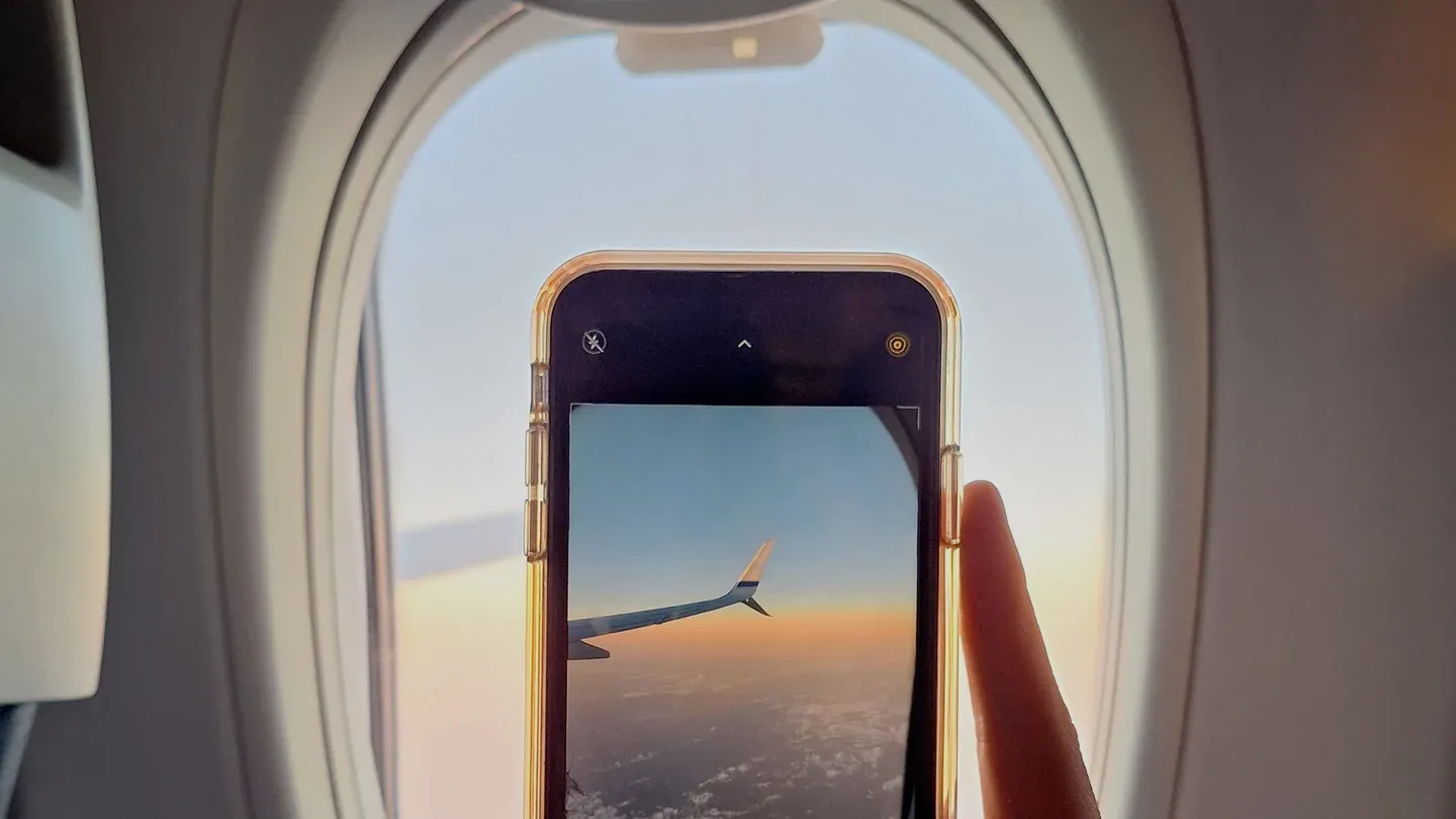 A traveler holding a smartphone in front of an airplane window with the wing visible