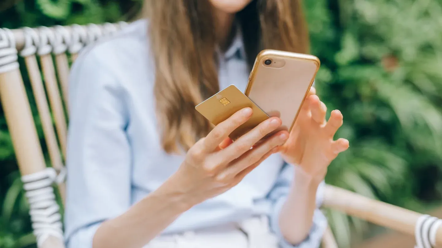 A person sitting outdoors holding a smartphone in one hand and a payment card in the other